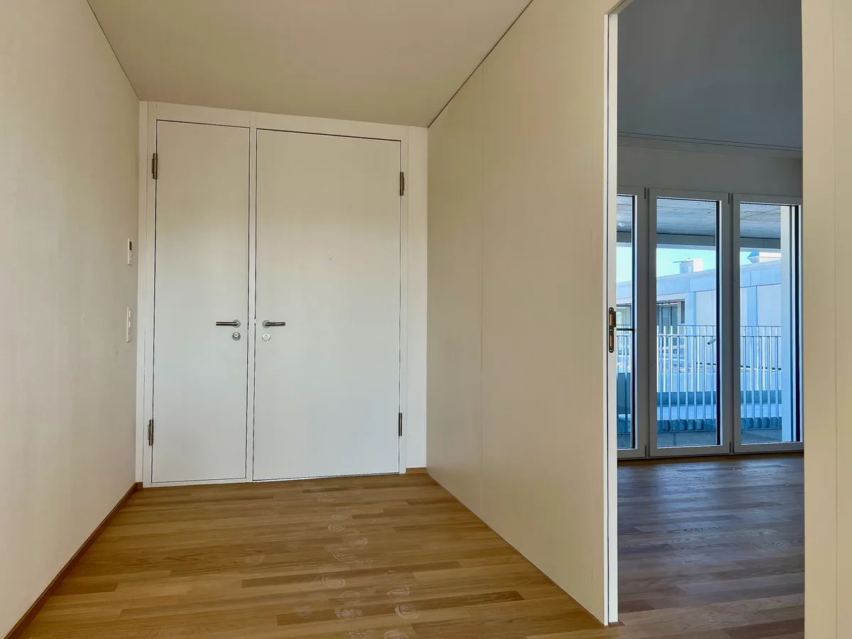 Man sieht links einen breiten Gang, an dessen Ende eine doppelte Tür verbaut ist. Rechts wird der Blick auf ein weiteres Zimmer freigegeben, das auch über eine bodenlange Fensterfront verfügt.